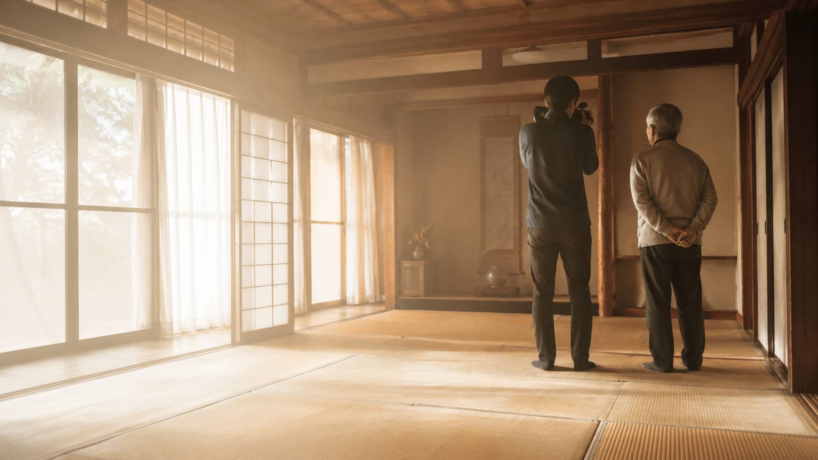 障子から光の差す畳の部屋で、撮影者がビデオカメラを構えて家の中を撮っている。その少し後ろに家の持ち主が立ち、同じ方向を見ている。ふたりとも後ろ姿。
